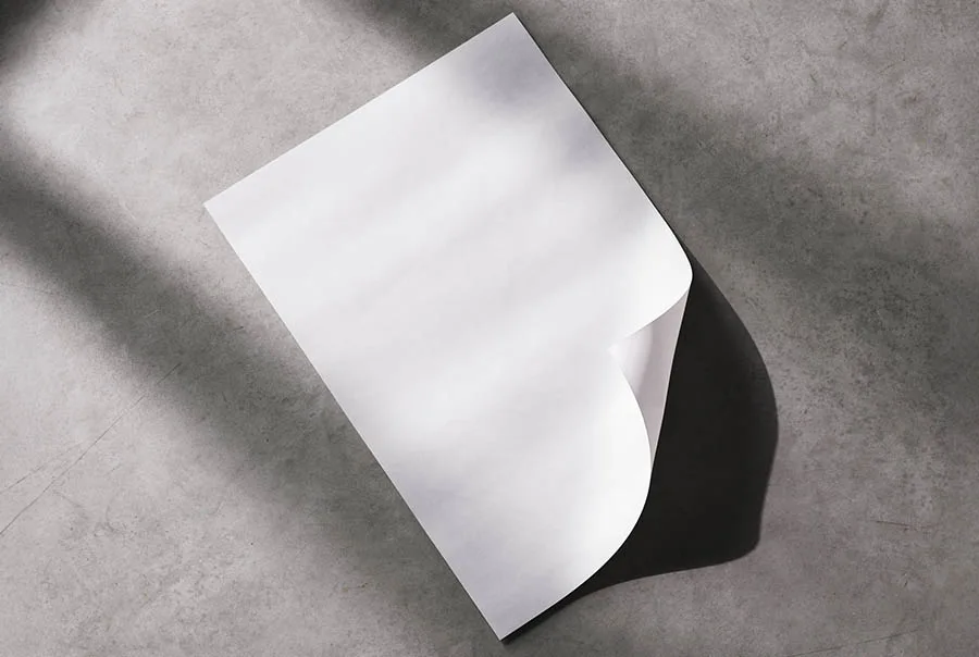 White poster with a curled corner casting a shadow on a textured gray concrete floor, viewed from above.