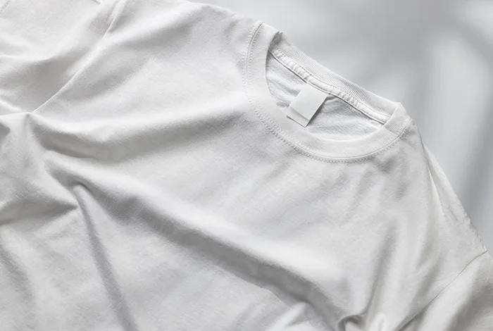 Close overhead view of a plain white crew-neck T-shirt laid flat on a light gray surface.