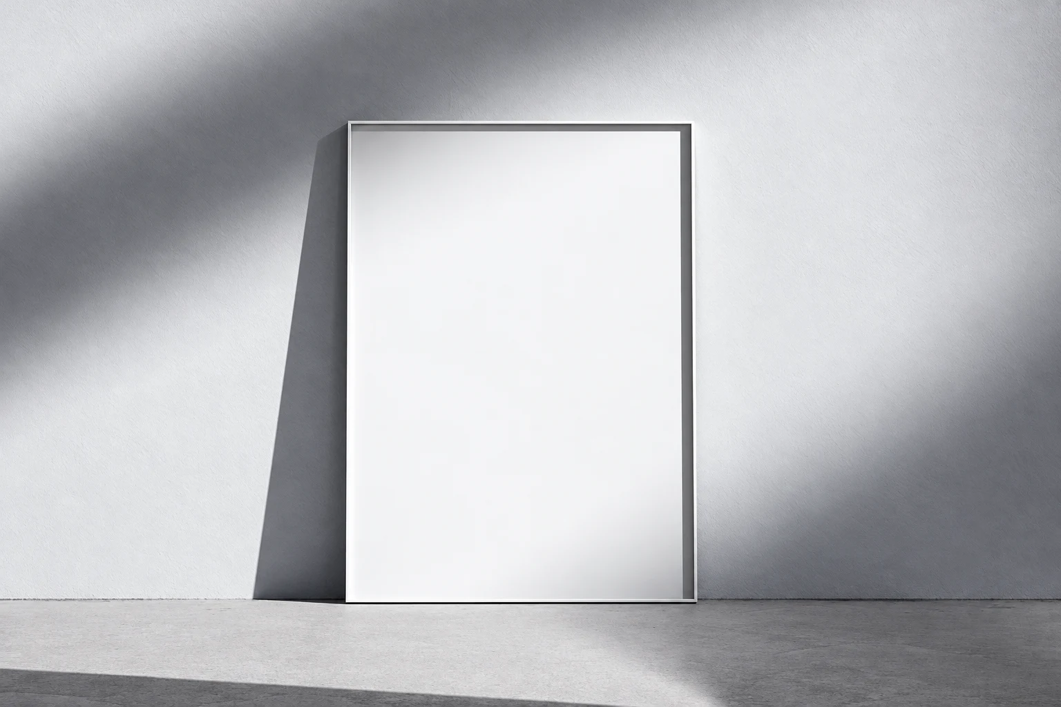 Blank portrait poster with a narrow dark frame leaning against a pale gray wall on a gray floor, lit from the side.