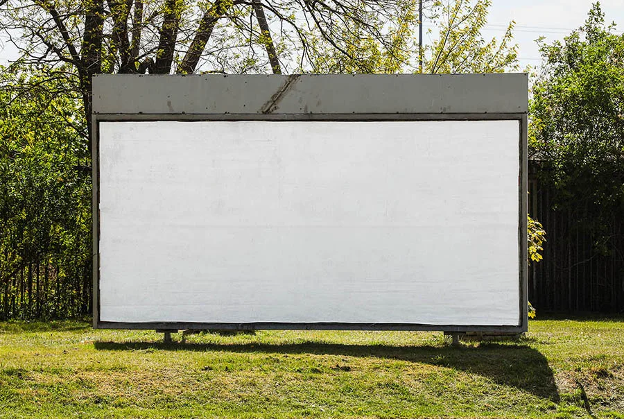 Straight-on view of a blank white billboard in a weathered gray frame, set in a grassy clearing with trees behind it.