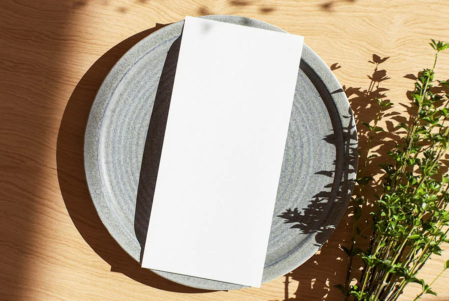 A blank white DL menu card lying across a grey stoneware plate on a wooden table with leafy shadows.
