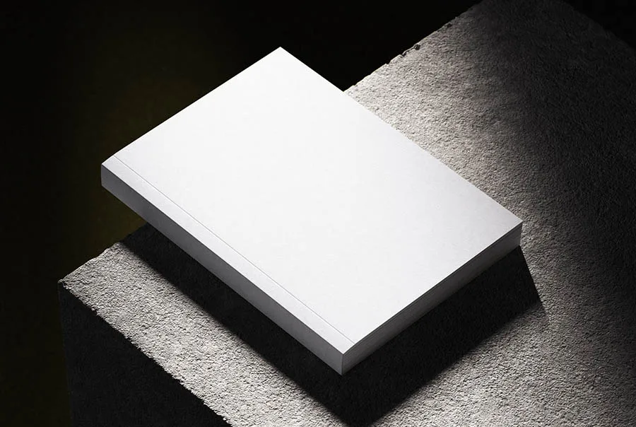 Closed blank white softcover book viewed from above at an angle on rough concrete, with strong side lighting and a dark background.