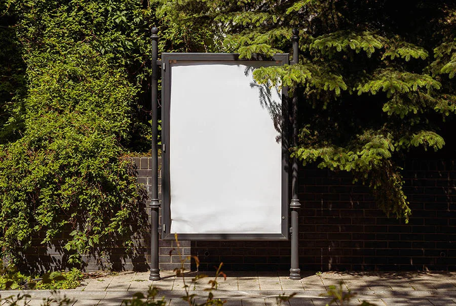 Blank white citylight poster in a dark metal frame on a leafy street beside a brick wall.