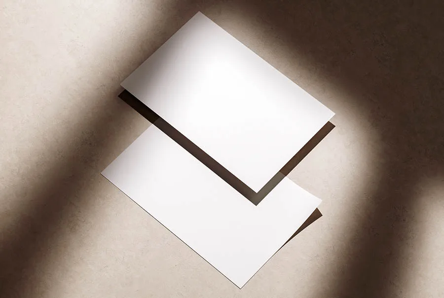 Two blank white flyers overlap on a textured beige surface in strong sunlight, viewed from above.