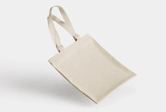 Plain natural-canvas tote bag at an angle against a pale gray background, with its handles raised behind the blank front panel.